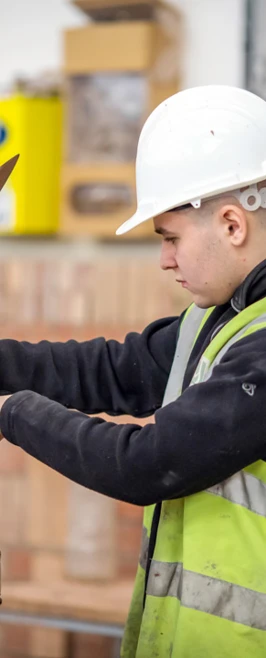 A bricklaying student in protective gear and high-visibility vest concentrating while applying mortar with a trowel to build a brick wall. A bricklaying student in protective gear and high-visibility vest concentrating while applying mortar with a trowel to build a brick wall.
